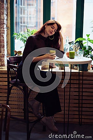 Beautiful serious stylish fashionable smart girl is sitting in cafe and drinking healthy smoothie or latte vegan. Charming Stock Photo