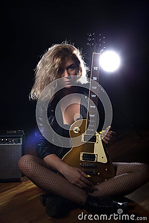 A beautiful and blonde is sitting on a wooden floor and holding an electric guitar. Rear contour light and glare. Stock Photo