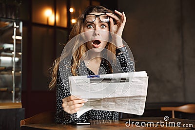 Beautiful shocked excited young woman sitting in cafe indoors reading newspaper Stock Photo