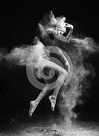 Beautiful slim girl wearing a gymnastic bodysuit covered with clouds of the flying white powder jumps dancing on a dark. Artistic Stock Photo