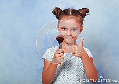 Beautiful small kid girl applying tonal cosmetic brush and showing thumb up sign on blue background with empty copy space Stock Photo