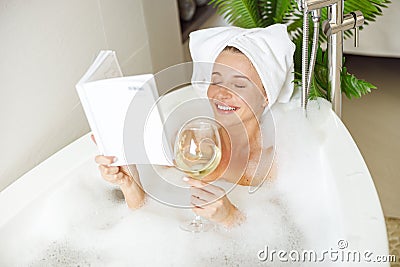 Beautiful smiled young woman lying in bath with foam, reading book and drinking wine. Stock Photo