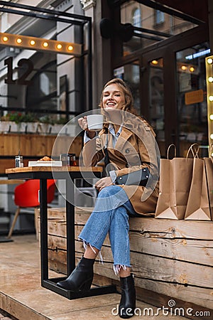 Beautiful smiling girl in trench coat and jeans joyfully looking in camera holding white cup of tea in hand while Stock Photo