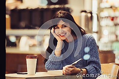 Beautiful smiling student woman reading a book in the cafe with warm cozy interior and drinking coffee Stock Photo