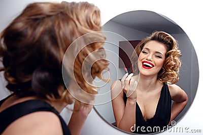 Beautiful smiling young woman with perfect make up, red lips, retro hairstyle in black dress, looking in mirror Stock Photo