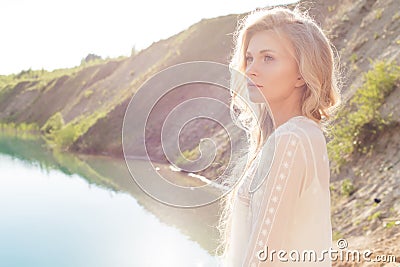 Beautiful soft bright girl with blond wavy hair stands on the shore of the lake at sunset on a bright sunny day Stock Photo