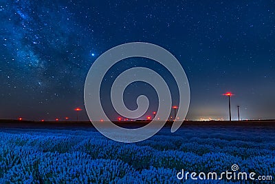 Beautiful starry night sky with milky way and blue sky over a field of lavender and red lights of wind turbines Stock Photo