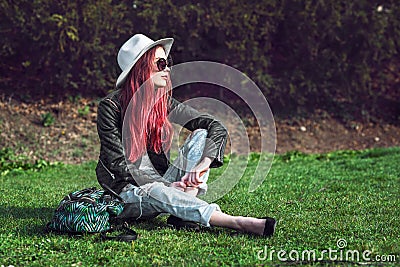Beautiful stylish red haired fashion hipster model woman sitting outdoors on green grass at park wearing sunglasses, hat and black Stock Photo