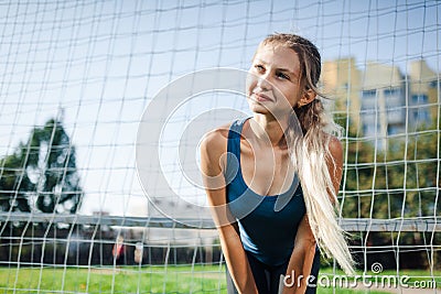Beautiful stylish woman in a blue shirt and leggings stands near a football goal at the stadium at sunset. Beautiful sunlight. A g Stock Photo
