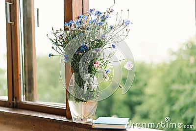 A bouquet of cornflowers and a book on the windowsill in a cozy home. Stock Photo