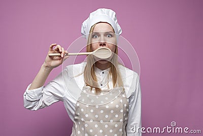 Beautiful surprised girl cook in kitchen clothes with a spoon closes her mouth on a pink background, woman housewife with kitchen Stock Photo