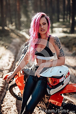 Modern tattooed hipster girl with bright pink hair sitting on her motocross bike in woods Stock Photo