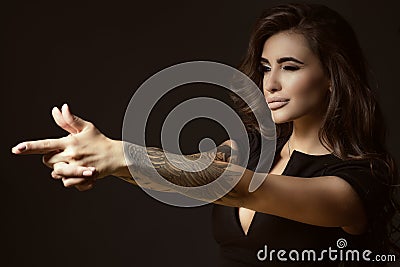 Beautiful tattooed woman with luxuriant shining wavy hair and perfect make up pretending to aim something with shooting gesture Stock Photo