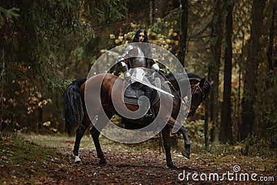 A beautiful warrior girl with a sword wearing chainmail and armor riding a horse in a mysterious forest. Stock Photo