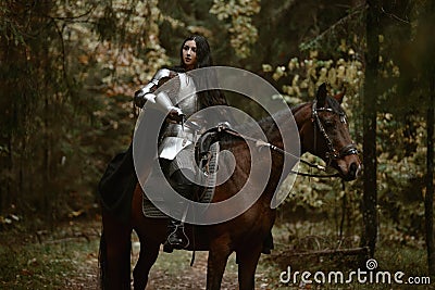 A beautiful warrior girl with a sword wearing chainmail and armor riding a horse in a mysterious forest. Stock Photo