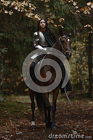 A beautiful warrior girl with a sword wearing chainmail and armor riding a horse in a mysterious forest. Stock Photo
