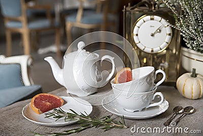 Beautiful white teapot, cups and saucer, antique clock, pumpkin, heather, rosemary and grapefruit. Still life. Stock Photo