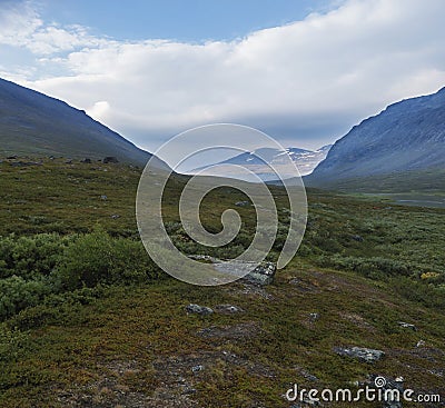 Beautiful wild Lapland nature landscape with blue glacial river, birch tree bushes, snow capped mountains and dramatic clouds. Stock Photo