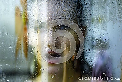 Beautiful woman behind the glass with water drops looking directly at camera. Girl takes a shower, voyeurism Stock Photo