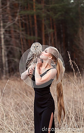 Beautiful woman in a black dress with an owl on his arm. Blonde with long hair in nature holding a owl. Romantic delicate girl Stock Photo