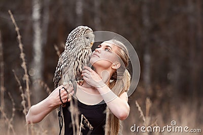 Beautiful woman in a black dress with an owl on his arm. Blonde with long hair in nature holding a owl. Romantic delicate girl Stock Photo