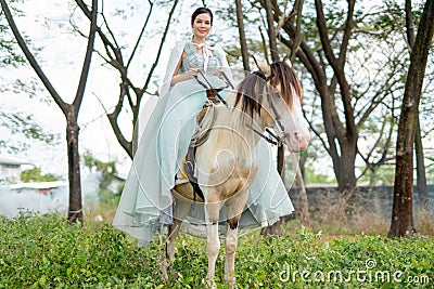 Beautiful woman with blue dress stay on white horse and stand in field near village during early morning also look at camera Stock Photo