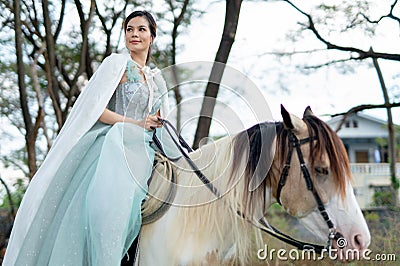 Beautiful woman with blue dress stay on white horse and stand in field near village during early morning Stock Photo