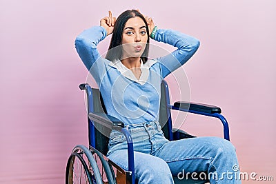 Beautiful woman with blue eyes sitting on wheelchair doing funny gesture with finger over head as bull horns Stock Photo