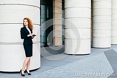 Beautiful woman with curly hair, slender legs, wearing black costume and high-heeled shoes, holding notebook in hands, arranging m Stock Photo