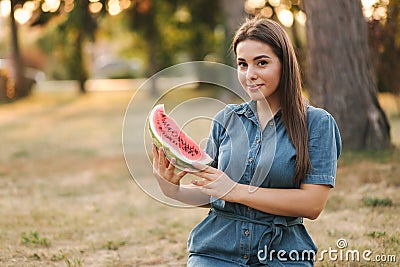 Beautiful woman in denimdress hold slice of watermelon in hands. Background of park Stock Photo