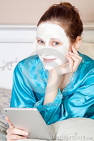 Beautiful woman with green nightie and white facial mask resting on bed and holding tablet before sleeping. Stock Photo