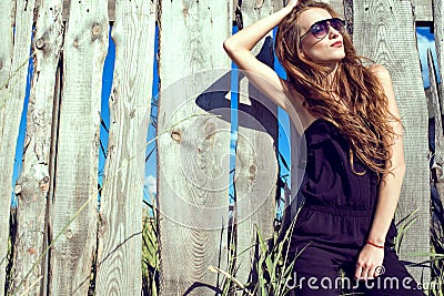Beautiful woman with long chestnut hair wearing jumpsuit and trendy aviator sunglasses at the wooden fence Stock Photo