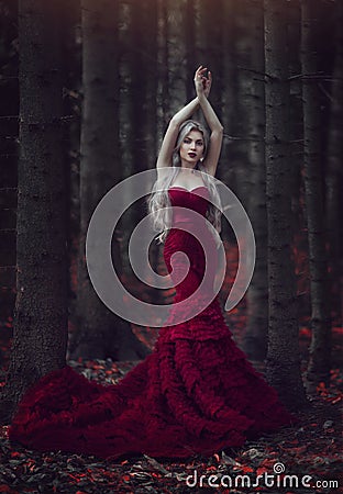 Beautiful woman with long white hair posing in a luxurious red dress with a long train standing in a autumn pine forest Stock Photo