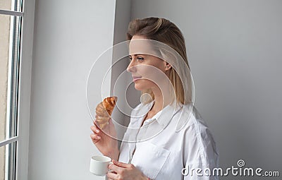 A beautiful woman looks out the window. Drink morning coffee with croissant Stock Photo