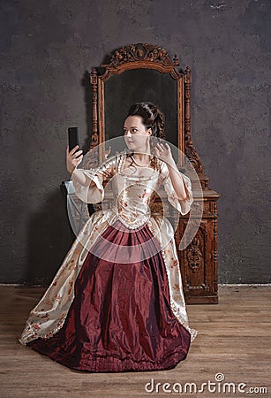 Beautiful woman in rococo style medieval dress sitting near console mirror table with mobile phone making selfie photo eclecticism Stock Photo