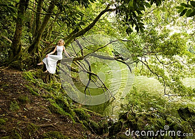 Beautiful woman sits in white long dress on branch of a tree, next to a lake Stock Photo