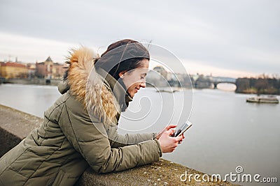 Beautiful woman standing by river in city Prague, text messaging. girl looking at phone on riverbank, with Prague bridge Stock Photo