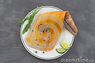 Beautiful yellow smoked haddock fish fillet on a plate with a branch of fresh arugula, lime and pink berry on grey slate Stock Photo