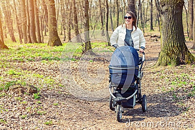 Beautiful young adult woman walking with baby in stroller through forest or park on bright sunny day. Healthy lifestyle and Stock Photo