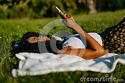 The beautiful young african girl with green eye shadows is laying on the grass and chatting, texting and browsing via Stock Photo