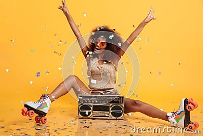 Beautiful young african woman with afro hairstyle throwing confetti, showing peace gesture while sitting in roller skates with bo Stock Photo