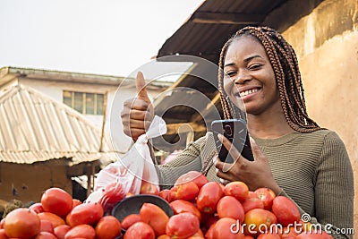 beautiful young african woman in a local african market using her smartphone giving thumbs up Stock Photo