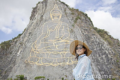 Beautiful young aisan woman is standing and smile on the background of mountain with golden statue of buddha on a hillside, Buddha Stock Photo