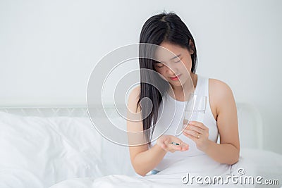 Beautiful young asian woman holding drug and glass of water while ill sitting on bed in the bedroom at home. Stock Photo