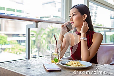 Beautiful young asian woman in red dress sitting in the restaurant looking out the window calling with smartphone . happy elegant Stock Photo