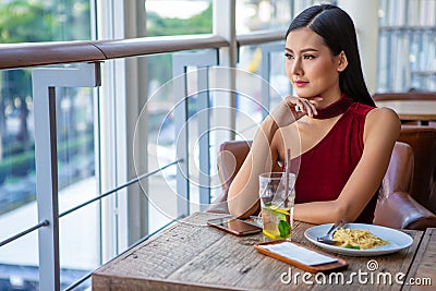 Beautiful young asian woman in red dress sitting in the restaurant looking out the window . happy elegant lady sitting on table Stock Photo