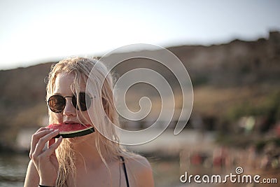 Beautiful young blonde female with sunglasses eating watermelon on the beach Stock Photo