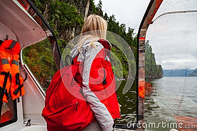 Beautiful young blonde girl in an orange life jacket standing on the edge and looking far in a motor boat during an excursion and Stock Photo