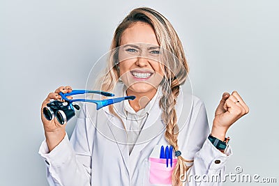 Beautiful young blonde woman holding scientist glasses screaming proud, celebrating victory and success very excited with raised Stock Photo