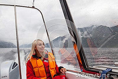 Beautiful young blonde woman in an orange life jacket sits in a motor boat during an excursion and sailing on Lake Teletskoye in Stock Photo